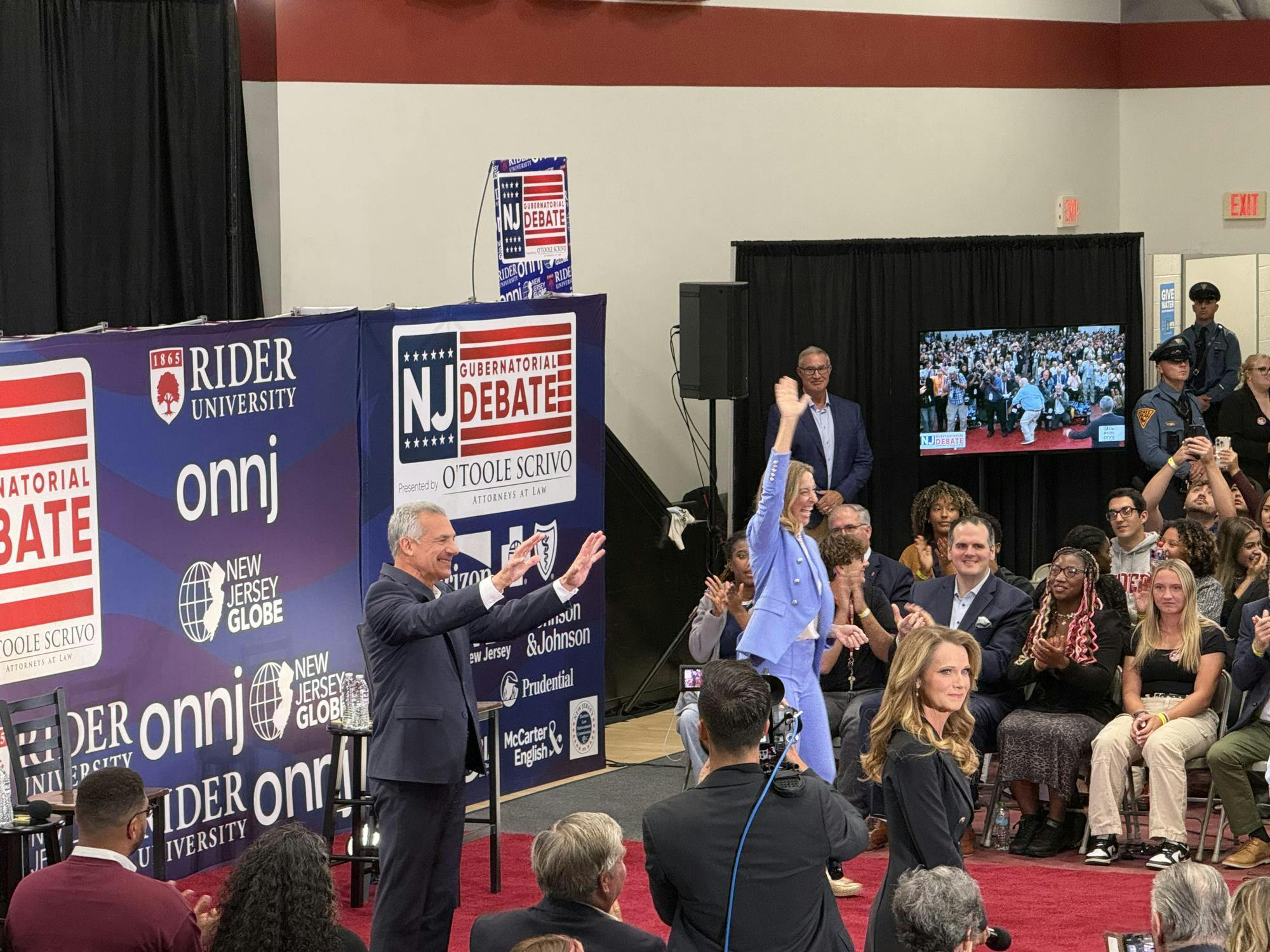Two people are standing in front of a wall with various logos for newspapers on it. They are waving at a large crowd.