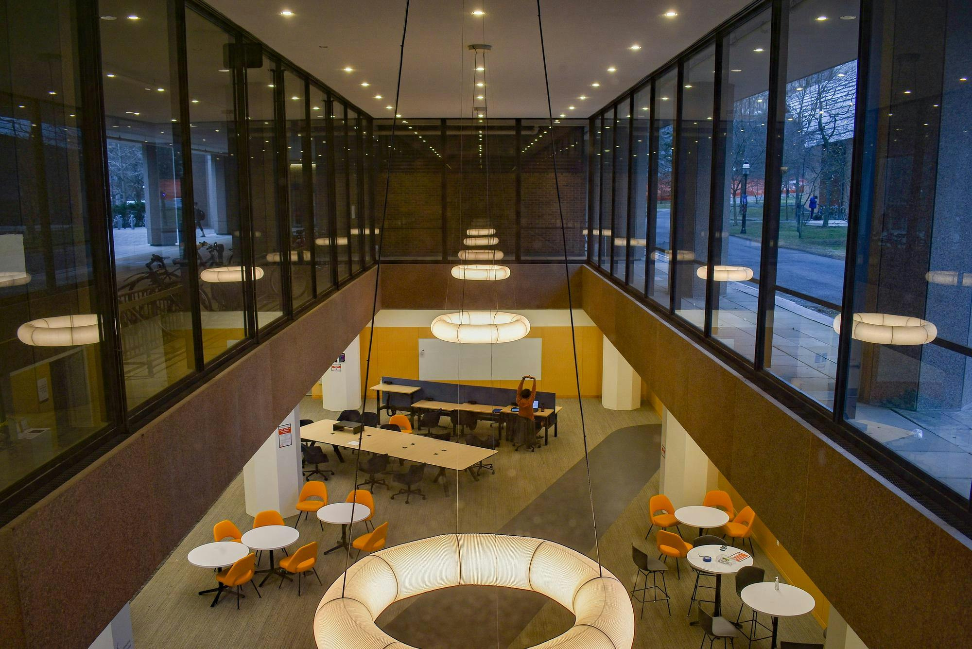 Three circular lights hang from the ceiling over a library, with neutral carpet floors, tables, and chairs.