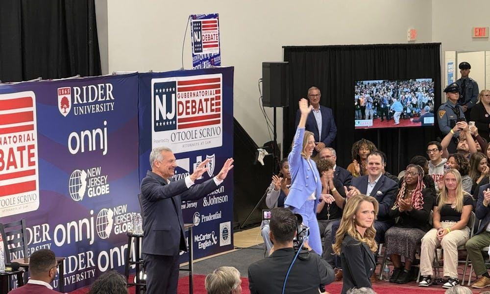 Dos personas están de pie frente a una pared con varios logotipos de periódicos. Saludan a una gran multitud.