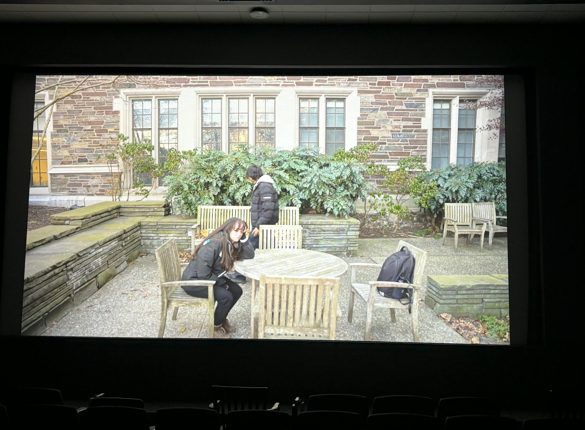 There is a beige gothic building with green plants and there is an outdoor brown, wooden table and chair. A light-skinned person with brown hair in a black jacket is sitting in one of the chairs. A person with dark hair and light-skin, also wearing a black jacket, is walking in the background.