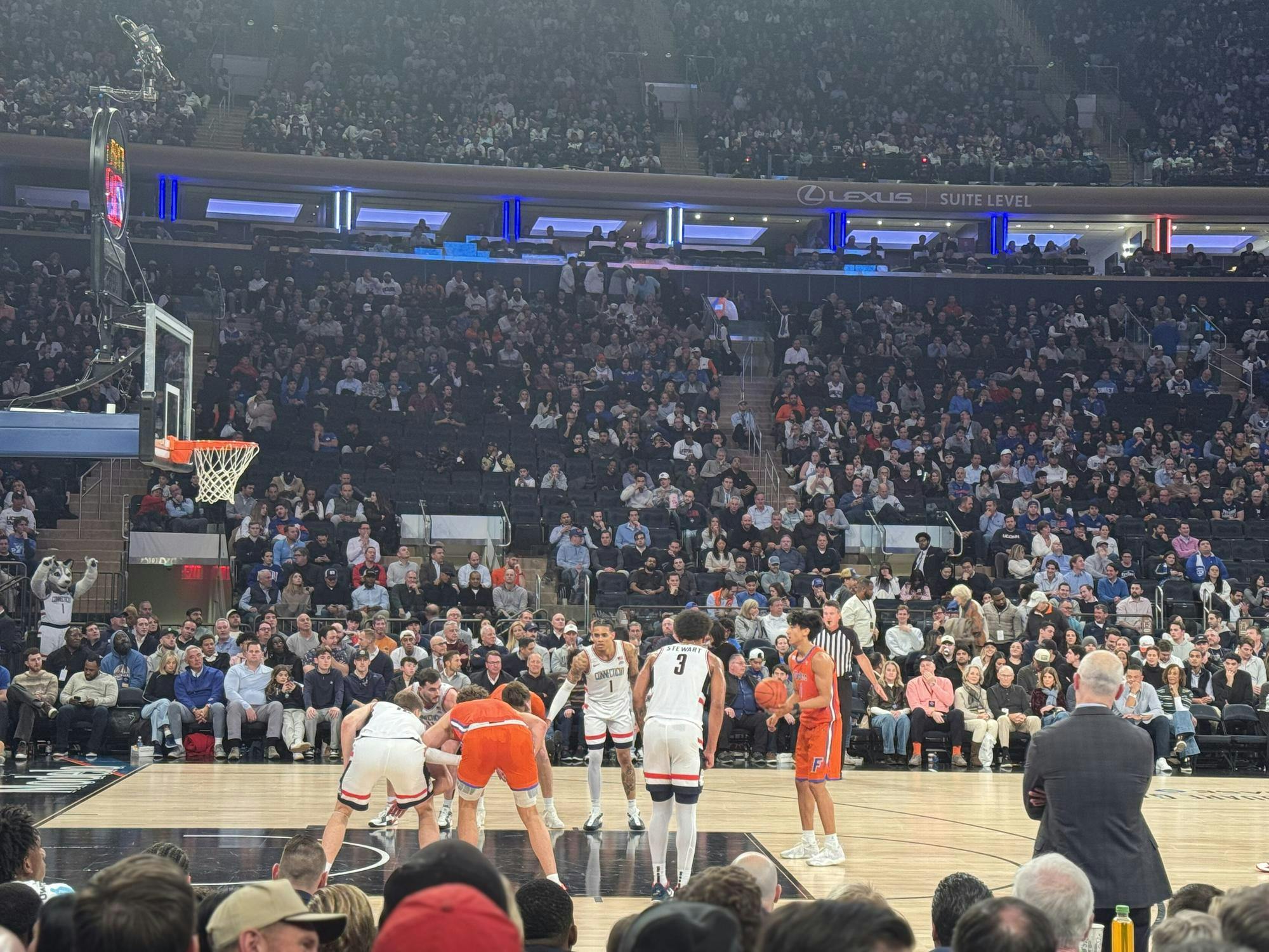 A basketball player prepares to shoot a free throw as players line up along the lane, with a referee and coach on the sideline and a packed arena crowd in the background.