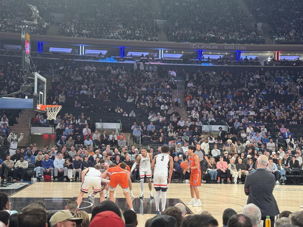 A basketball player prepares to shoot a free throw as players line up along the lane, with a referee and coach on the sideline and a packed arena crowd in the background.