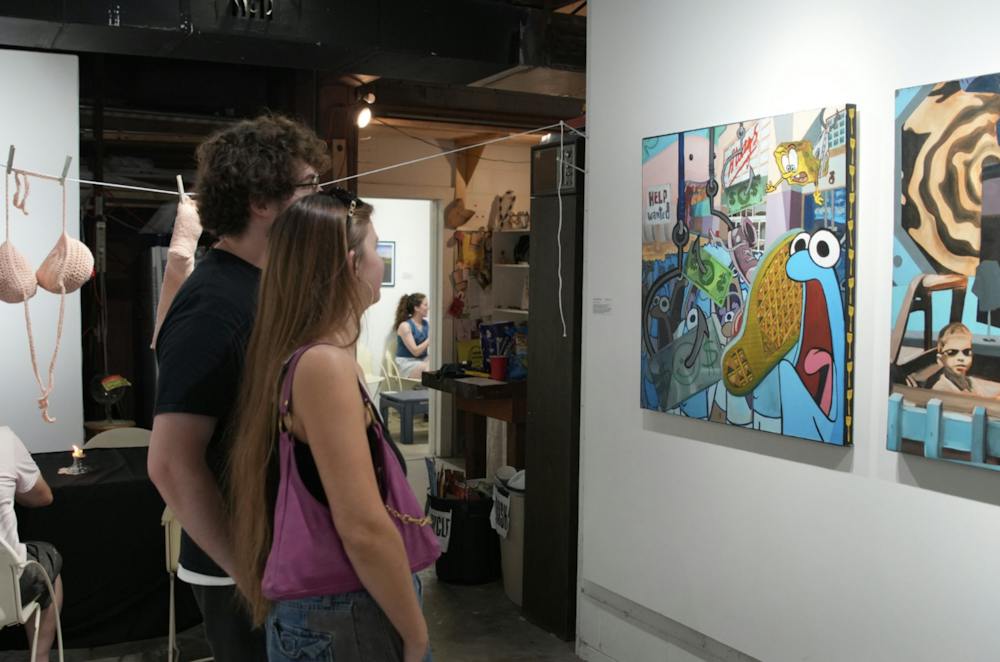 Two visitors take in paintings hung along the gallery wall at Storage Space Gallery. Crocheted soft sculptures hang from a clothesline at left.