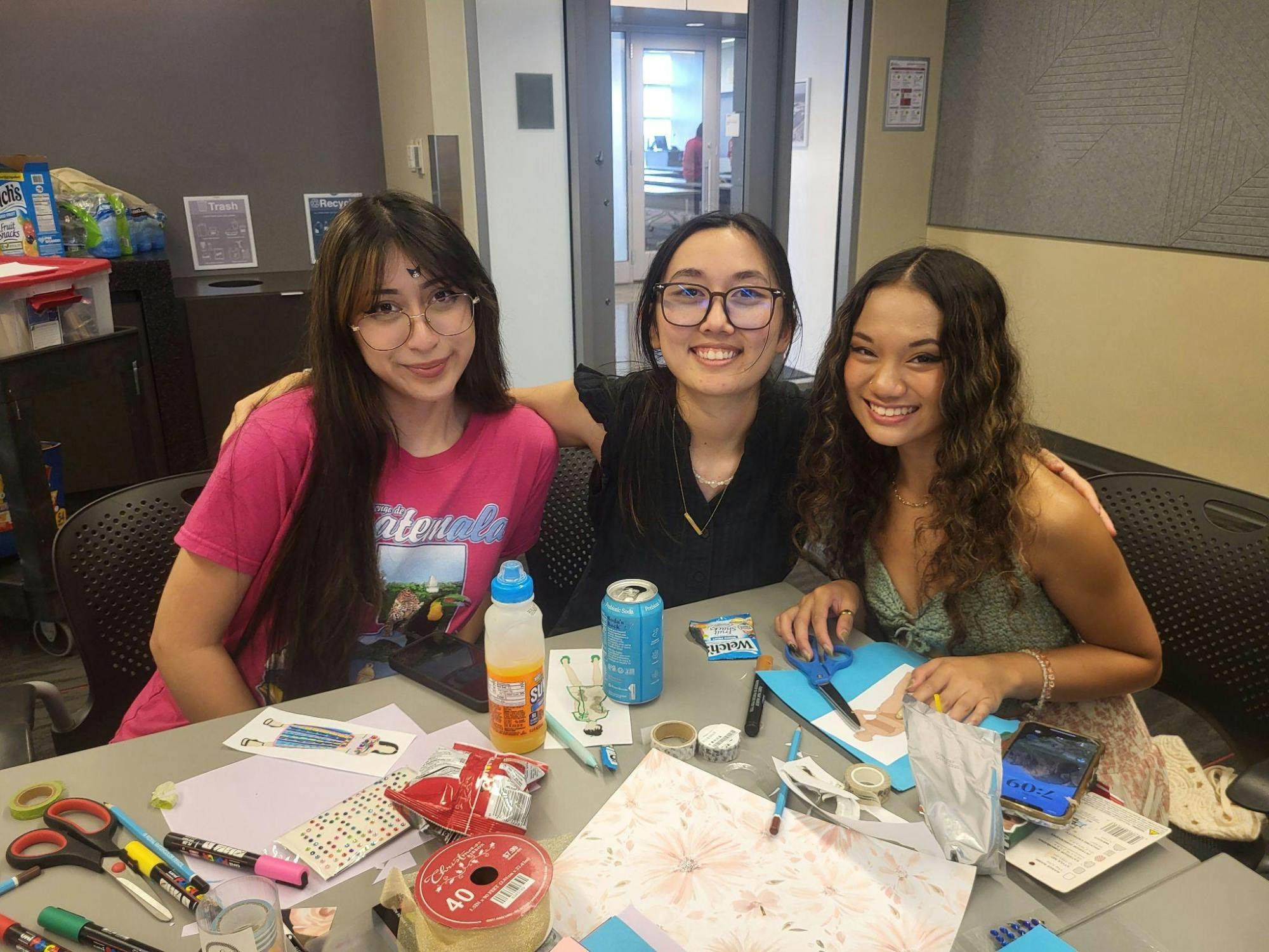 Three students smile at a table covered with craft supplies and snacks.