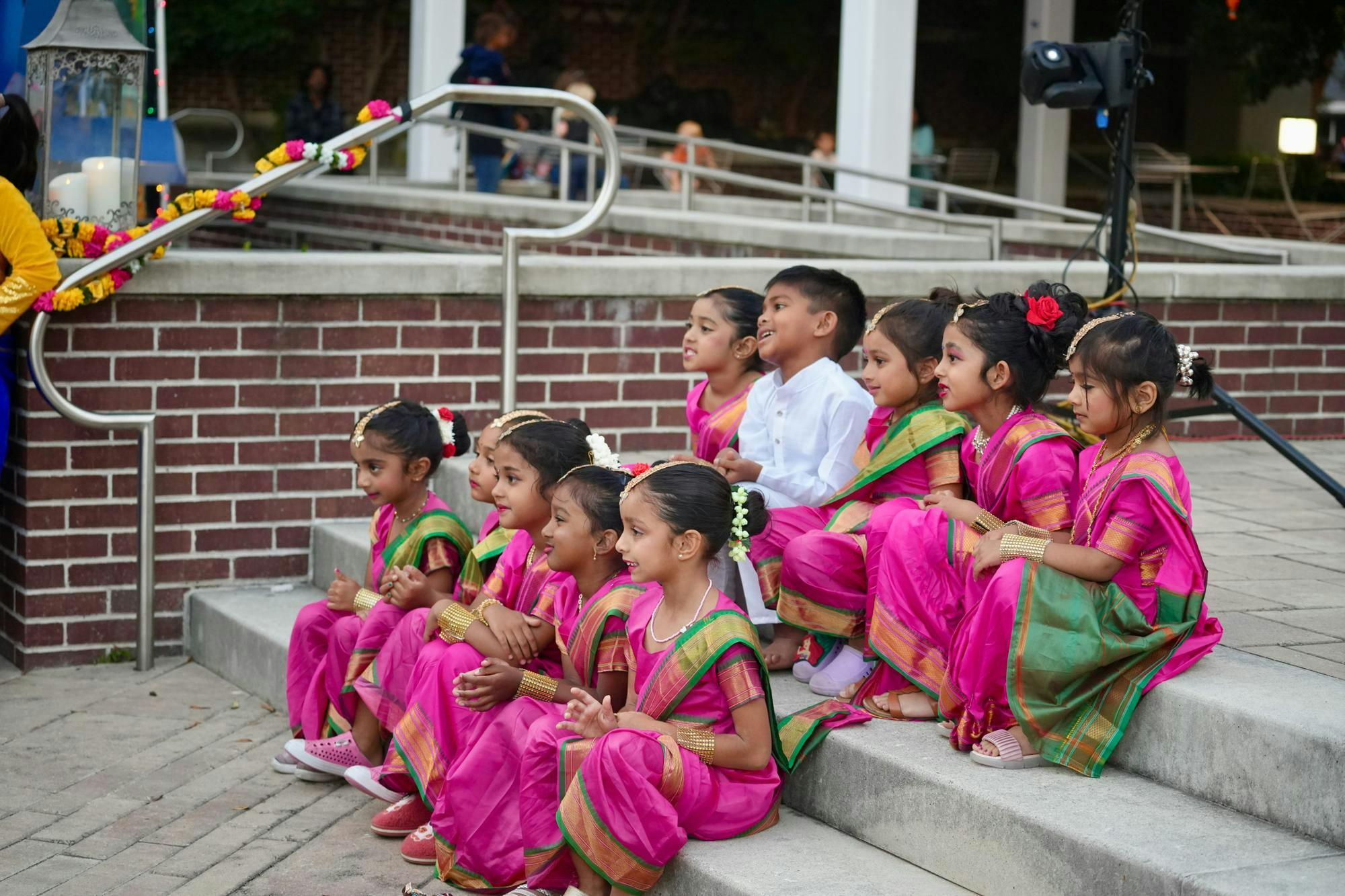 A group of children in bright magenta traditional Hindu outfits and one young boy in an all white outfit sit on concrete steps together while smiling for a picture. 