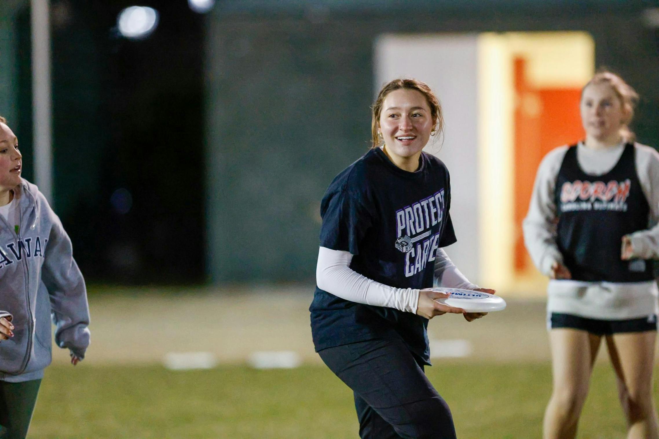 A young woman wearing a black graphic t-shirt and white long sleeves underneath holds a frisbee while looking ahead with a focused yet happy expression. She appears to be engaged in a nighttime ultimate frisbee game, with two other players in the background—one wearing a gray "HAWAII" sweatshirt and the other in a black and white sports vest.