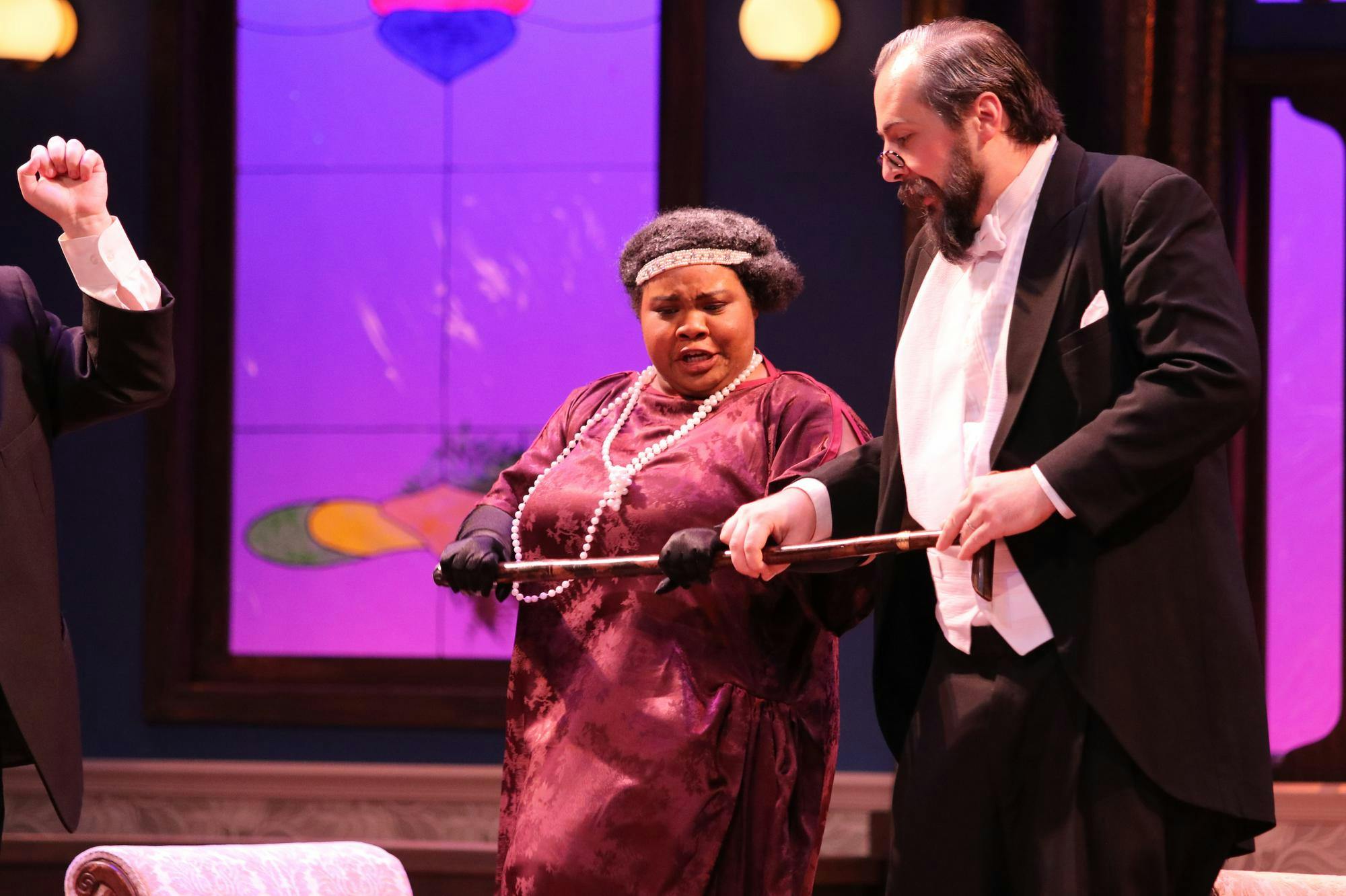 A woman wearing a velvet dress and a long pearl necklace stands next to a man wearing a black suit and white shirt. They are both holding a cane.