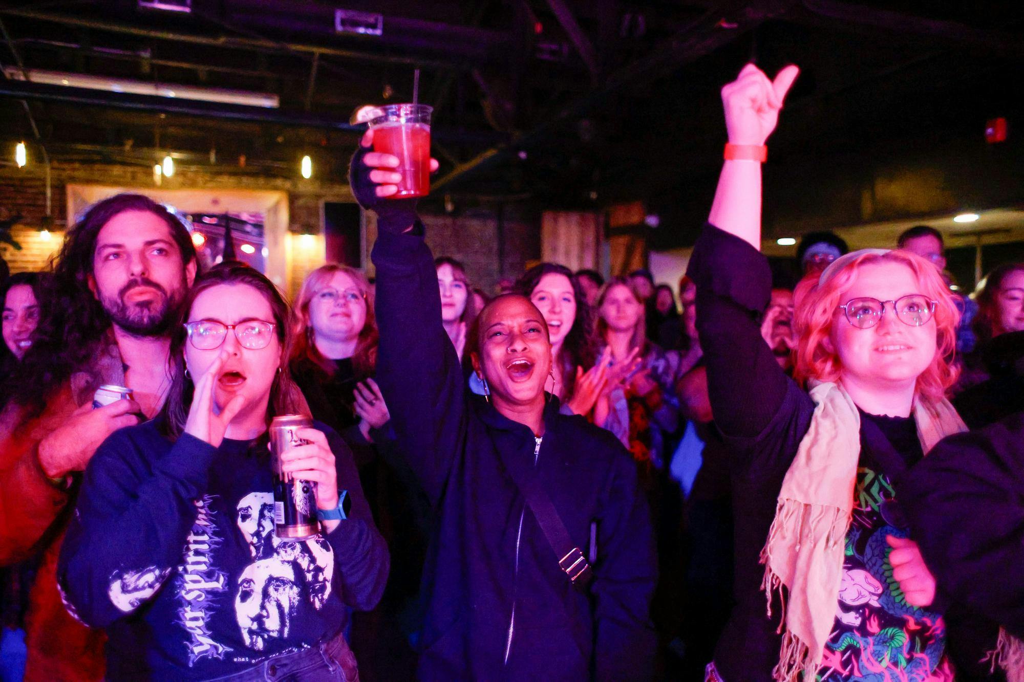 A vibrant crowd at a lively indoor event, with attendees expressing excitement and energy. In the foreground, a woman raises a drink in celebration, while another cheers enthusiastically. A mix of individuals, some holding drinks and others clapping or smiling, are illuminated by colorful event lighting. The background reveals the industrial-style venue with exposed beams and warm ambient lights, enhancing the festive atmosphere.