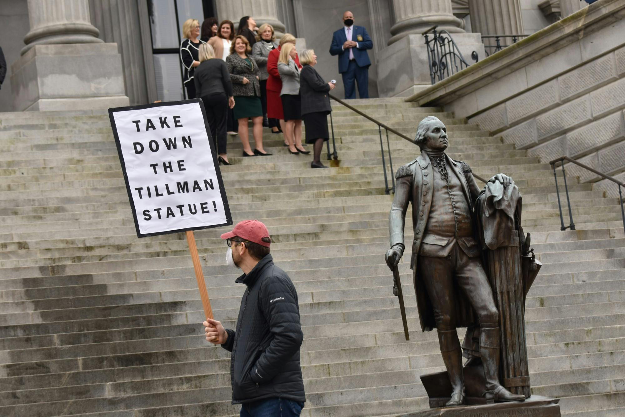A protestor holding a “Take Down the Tillman Statue” sign walks in front of the South Carolina Statehouse while Statehouse workers observe.