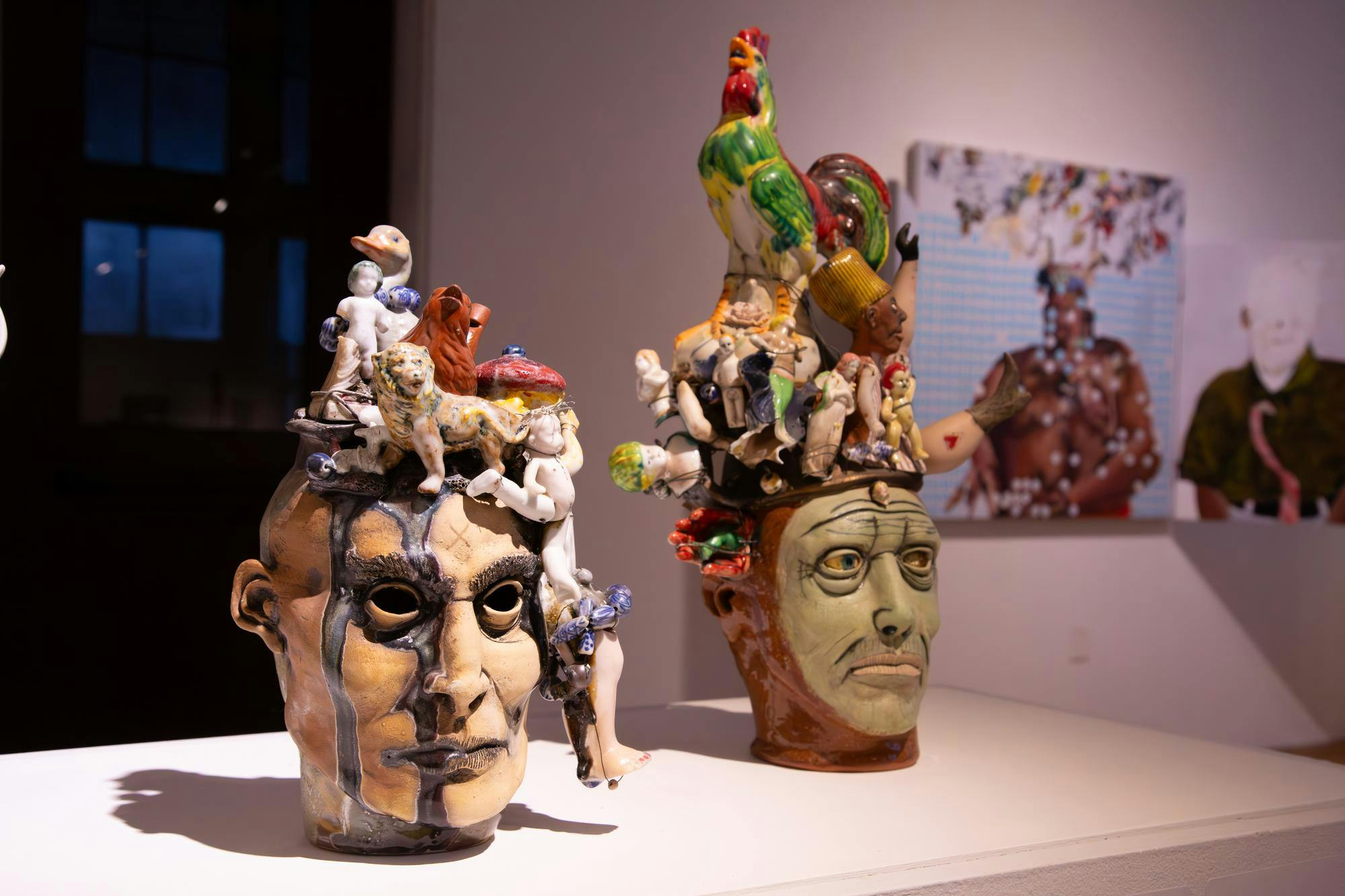 Two small sculptures sit next to each other on a white display table. The sculptures are both human heads with various miniature animals and people sitting on top of them.  