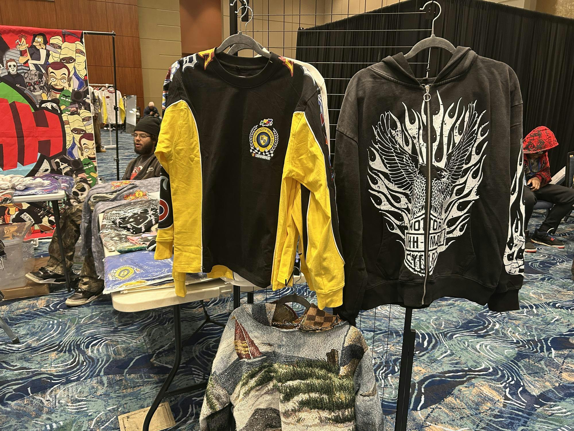 Two graphic hoodies hang on a display rack at an indoor streetwear market. One is black with bright yellow sleeves and racing-style patches and logos, while the other is a dark zip-up hoodie featuring a large white flaming eagle or winged graphic on the front. Behind them, folded shirts and illustrated tapestries sit on tables as vendors and shoppers move through the convention hall.