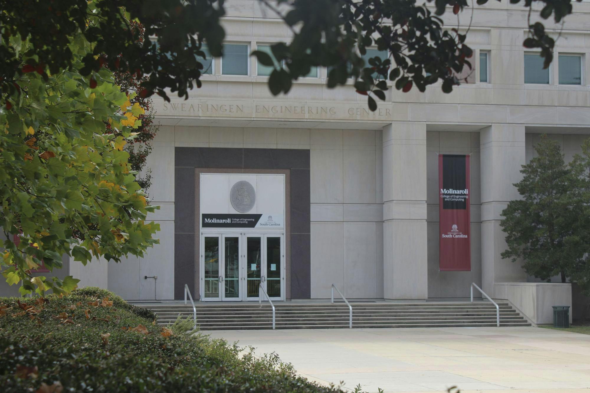 The front steps and doors of the Molinaroli College of Engineering appear behind branches of a tree. 