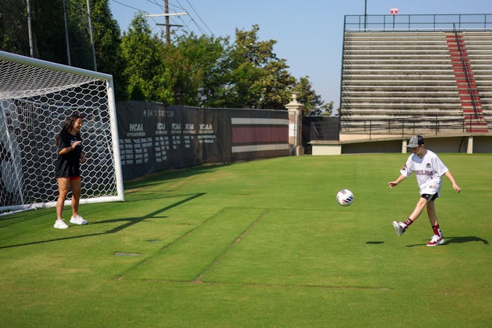 On a sunny soccer field, a young boy in a Gamecocks jersey kicks a soccer ball toward a goal while another person stands near the net, with empty bleachers and trees behind them.