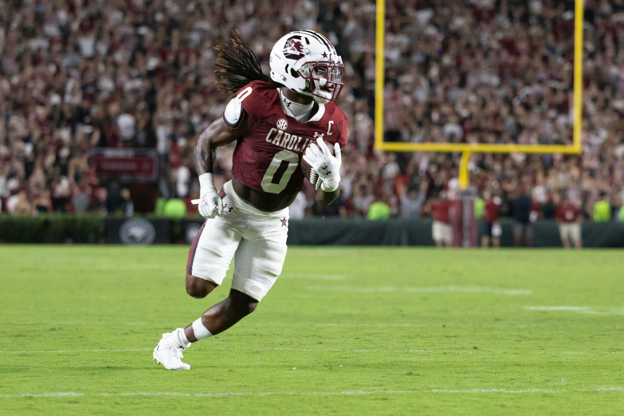 A University of South Carolina football player runs down the field holding a football in his right arm. The player is wearing a garnet jersey with the number 0 across the front and white shorts with a garnet and black stripe down the side. In the background, there is a yellow goalpost and crowds of people watching the game. 