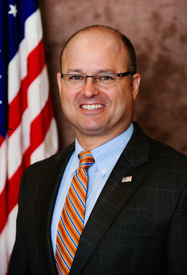 A man with glasses is pictured in front of a beige backdrop with an American flag behind his right shoulder. The man is wearing a dark gray suit, a light blue shirt and an orange, blue and white tie. 