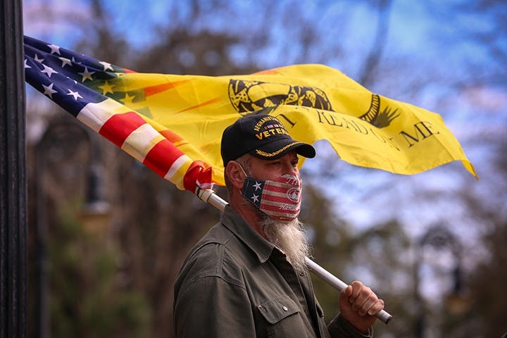 A protester holds a flag displaying a combination of the American and Gadsden flag while listening to a speaker.&nbsp;