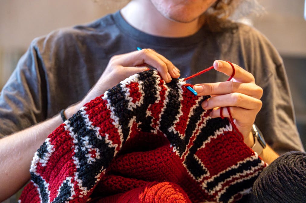 A person sits working on a crochet project made out of red, black and white yarn, with a blue crochet hook. 