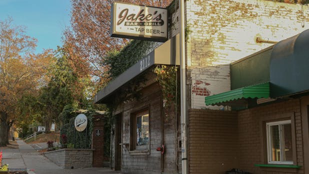 The outside of a brick building with a short black awning over its front side and a sign reading "Jake's Bar Grill Tap Beer" hanging from the front side.