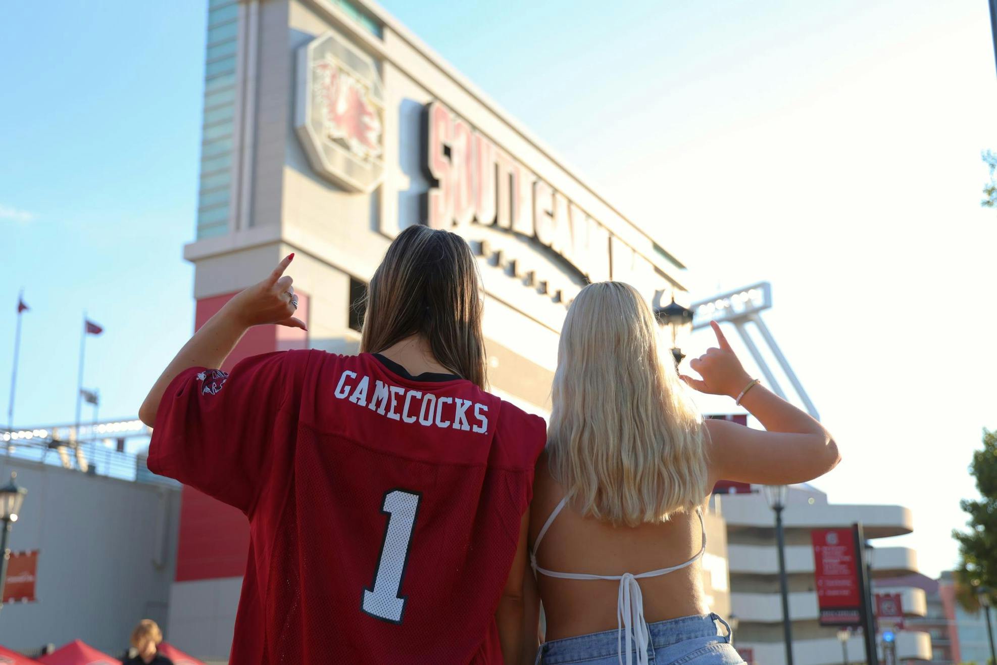 Two girls, wearing a red football jersey and a white top, look away from the camera towards Williams-Brice Stadium and hold up a "spurs up" hand gesture.