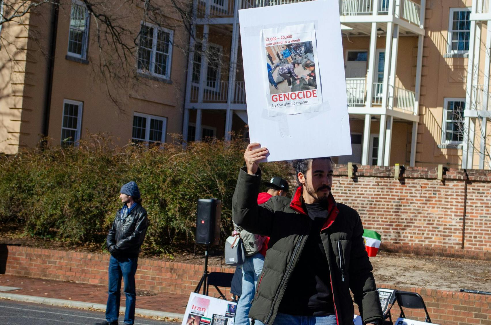 A man stands and holds a sign which accuses the Iranian government of murdering 12,000 to 20,000 people in a week, and accuses the Islamic regime of genocide.