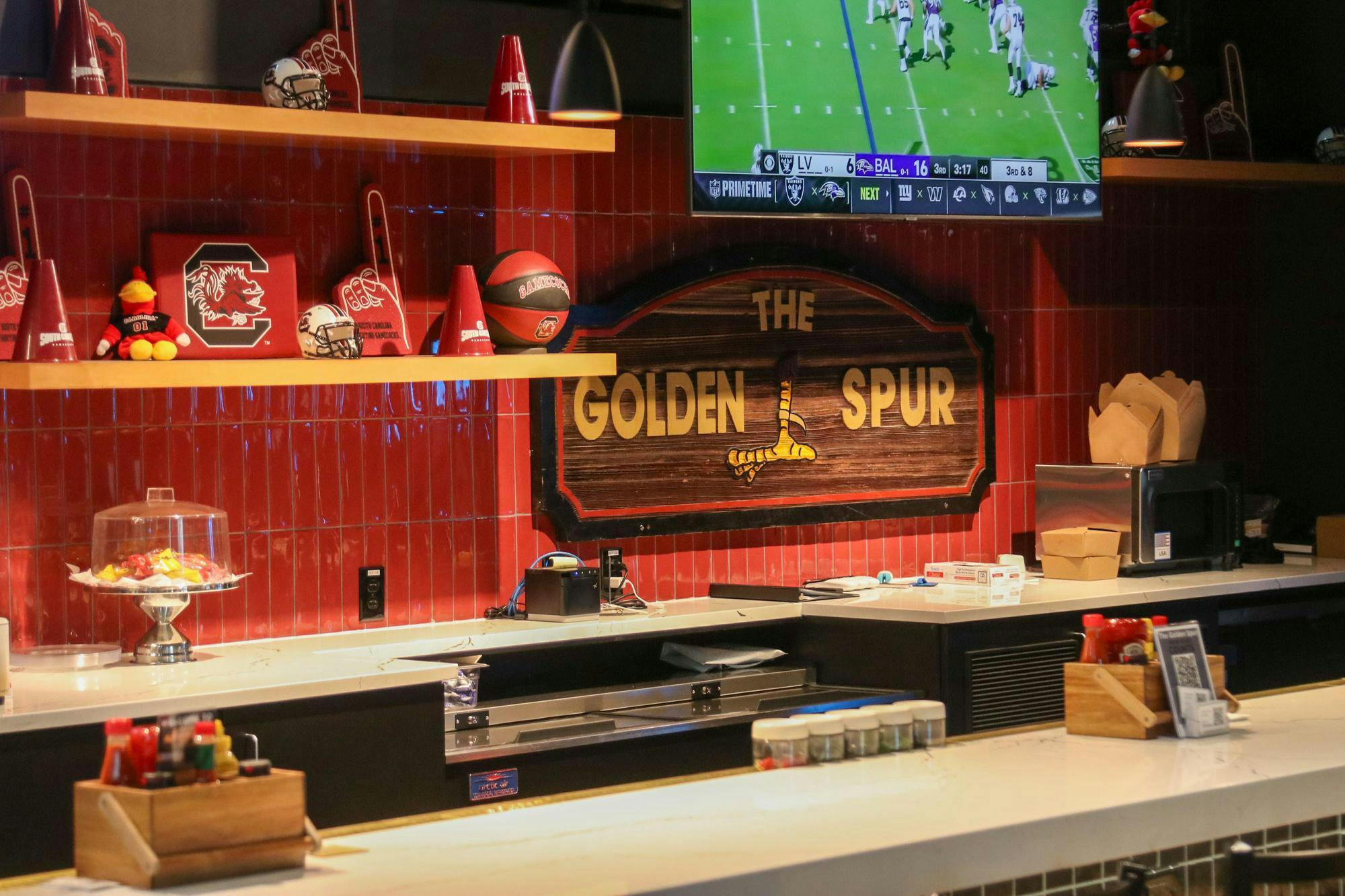 The inside of the Golden Spur with a bar in the foreground and a sign hanging behind the bar reading ‘The Golden Spur’ with a Gamecock foot in between it.