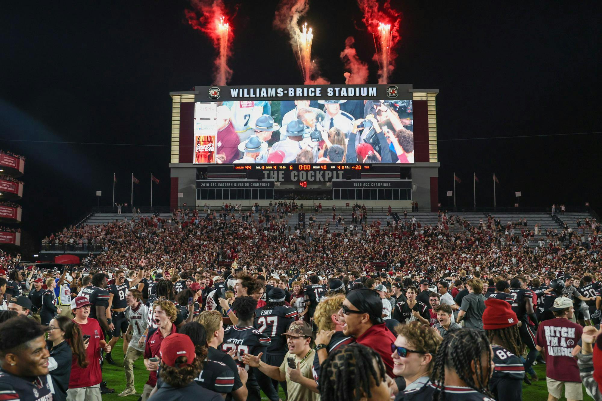 Football players wearing black jerseys and a mix of fans crowd the field of Williams-Brice Stadium. In the background, red and white fireworks shoot off from the top of the score board.