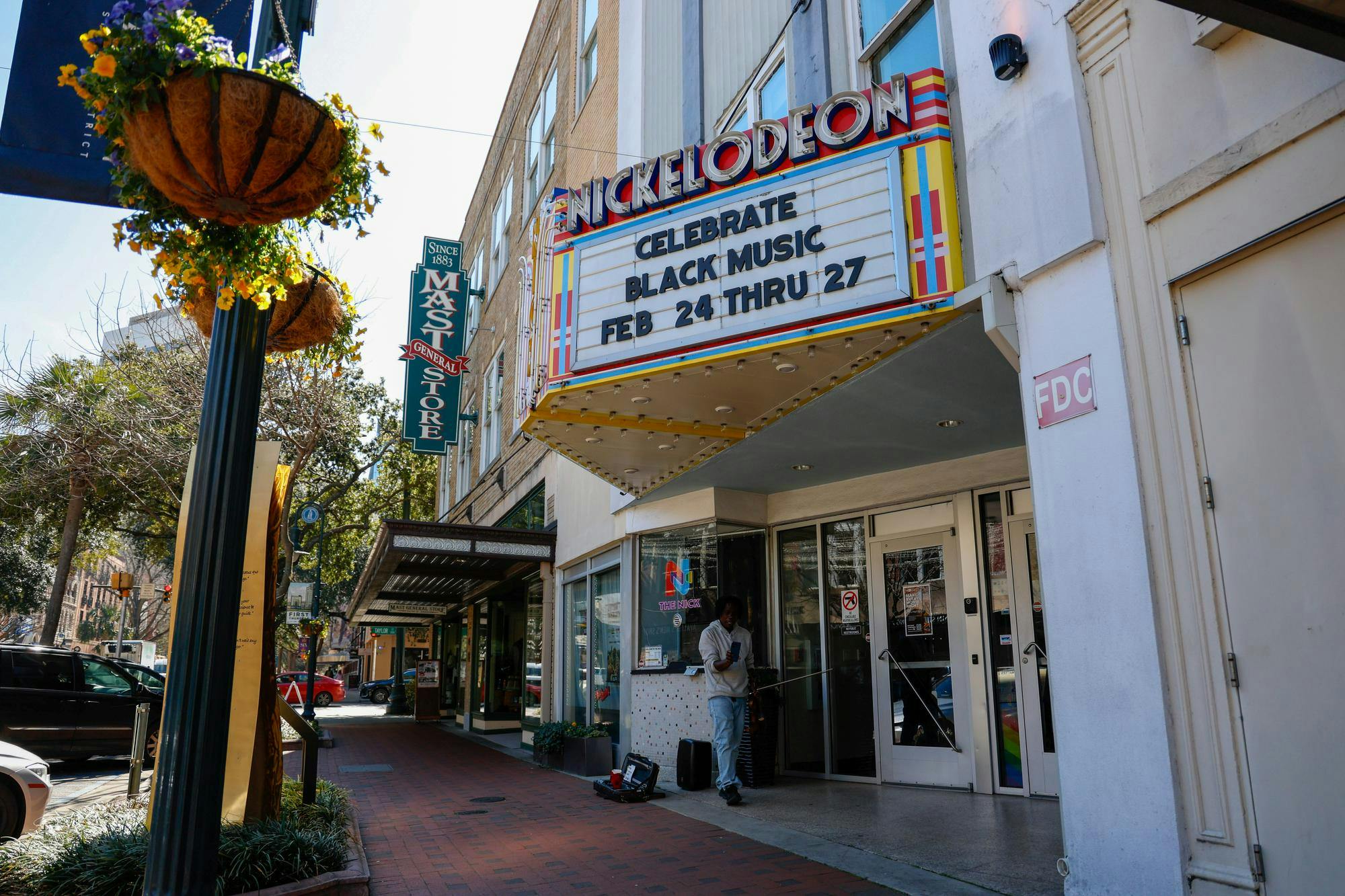 The image shows the marquee of the Nickelodeon Theatre, a historic movie theater with a retro-style sign. The marquee displays the message: "CELEBRATE BLACK MUSIC FEB 24 THRU 27." The sign has bold, metallic letters with a colorful, geometric border. To the left of the theater, there is a green sign for "Mast General Store," a historic retail chain. The background includes a brick building with large windows, and some tree branches are visible in the frame. A man walks below the sign, holding a violin in his hands.