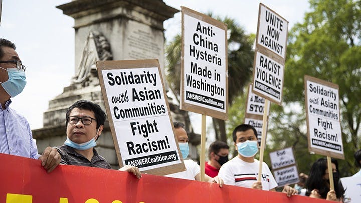 The crowd holds up a banner while waving signs that vary in message, all signs are about stopping racism, specifically racism against Asian communities.
