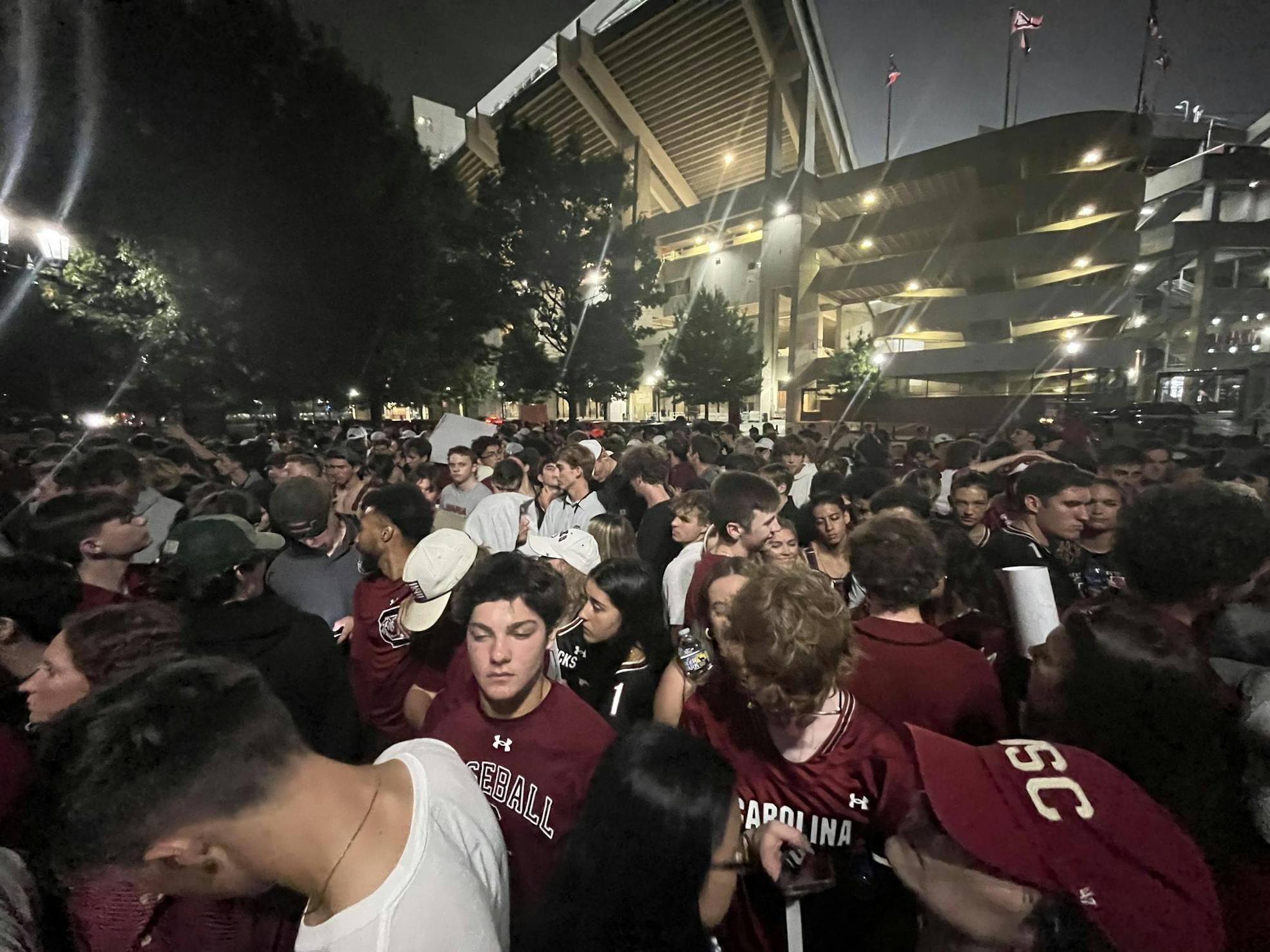 A large crowd of students wearing a variety of South Carolina gear stand shoulder to shoulder between Gamecock Park's gates and the street. A part of Williams-Brice Stadium can be seen in the background. 