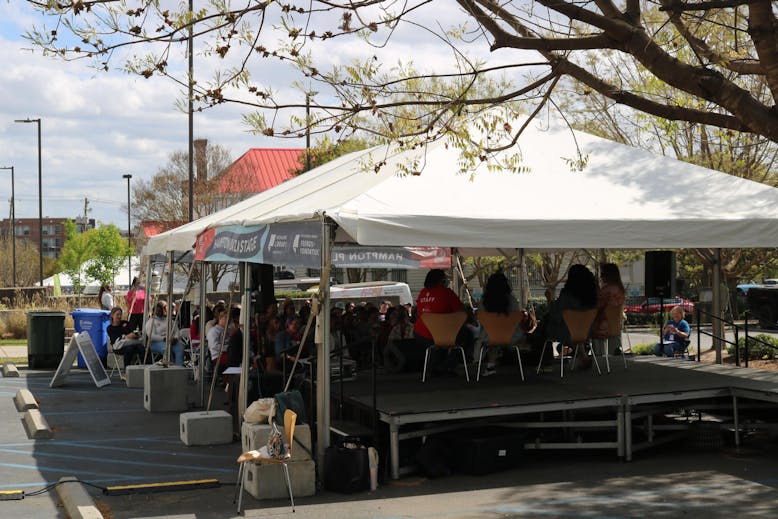 A white tent covers a group of people seated outdoors for a panel event, with four speakers seated on a stage in front of the crowd. 