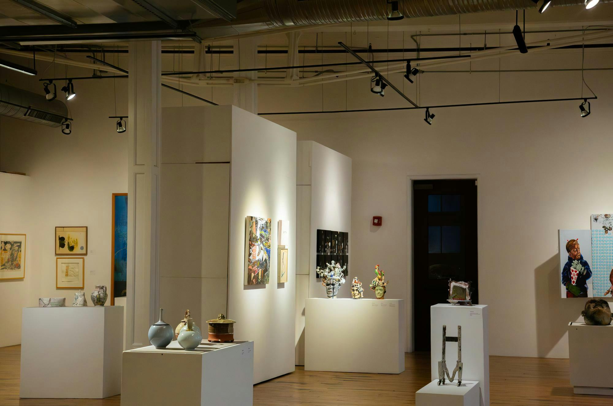 A wide view of an art gallery, the art displayed on all-white walls and tables. The art in the picture ranges from paintings to sculptures. 