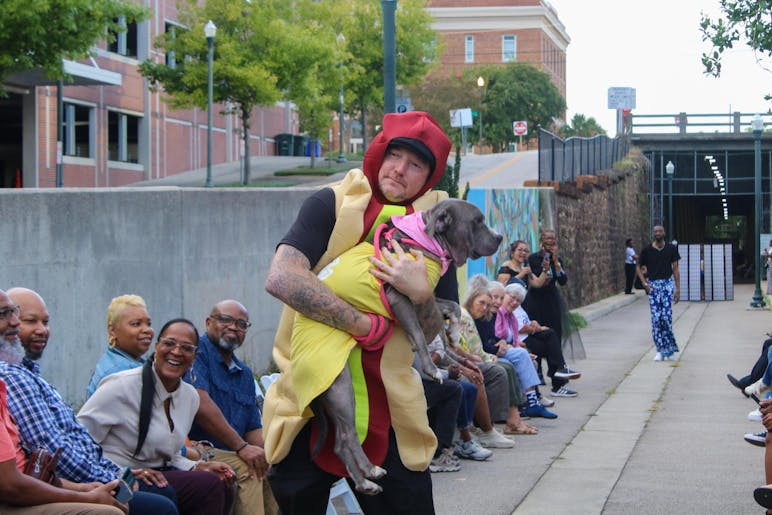 A man in a hot dog costume holds a dog in his arms as he poses at the end of a runway.