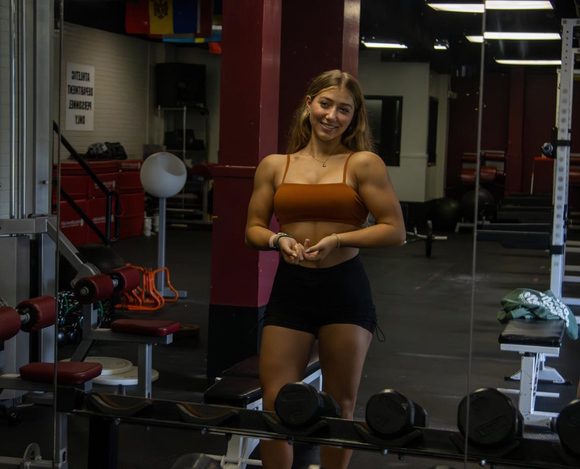 Jessica Dianda, in a brown and black gym outfit, is seen posing in a mirror in front of gym equipment. 