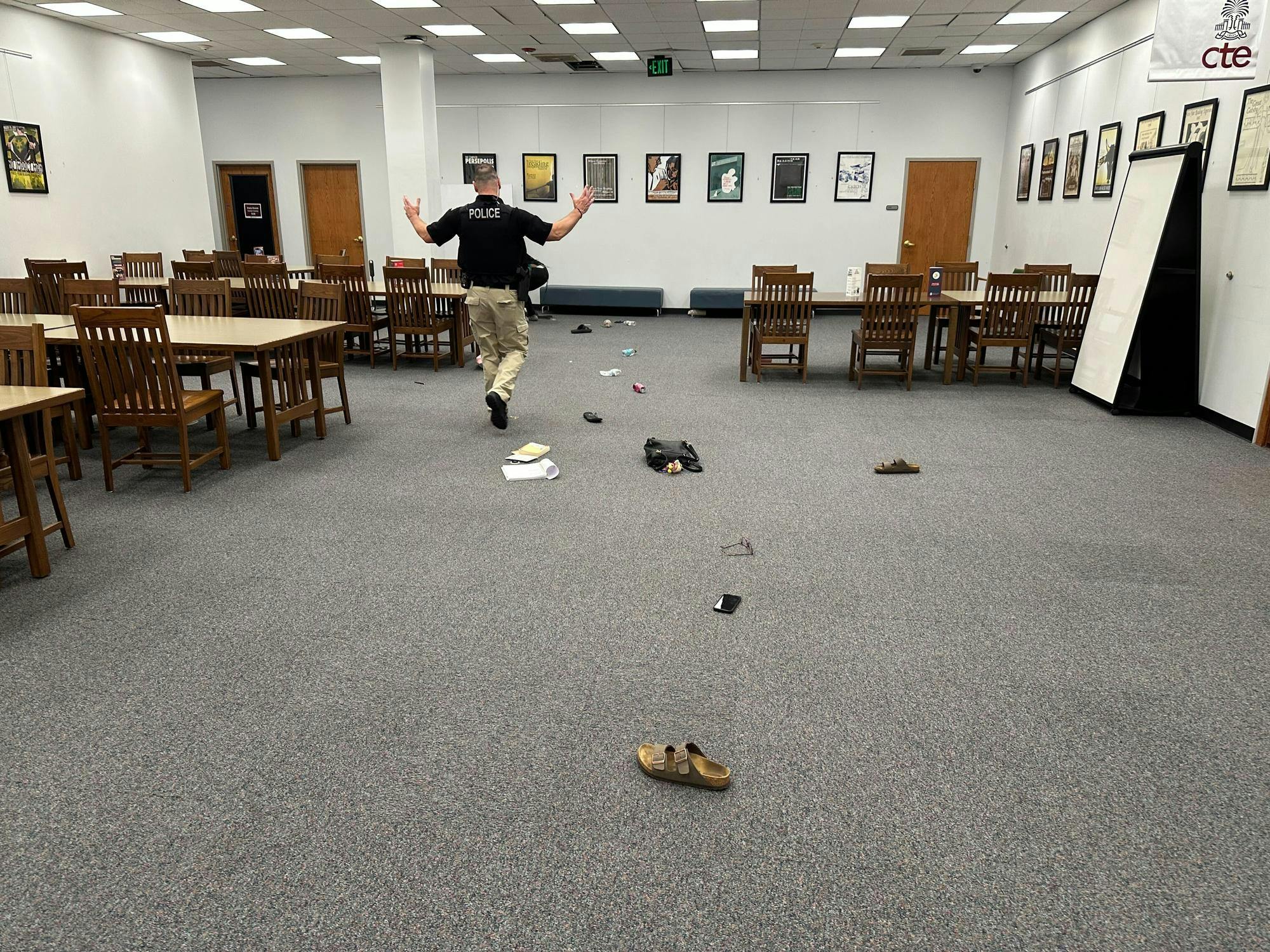 A police officer with “POLICE” printed on the back of his vest walks with arms raised inside a room lined with wooden tables and chairs. The floor is scattered with abandoned personal belongings including shoes, papers and a cell phone.