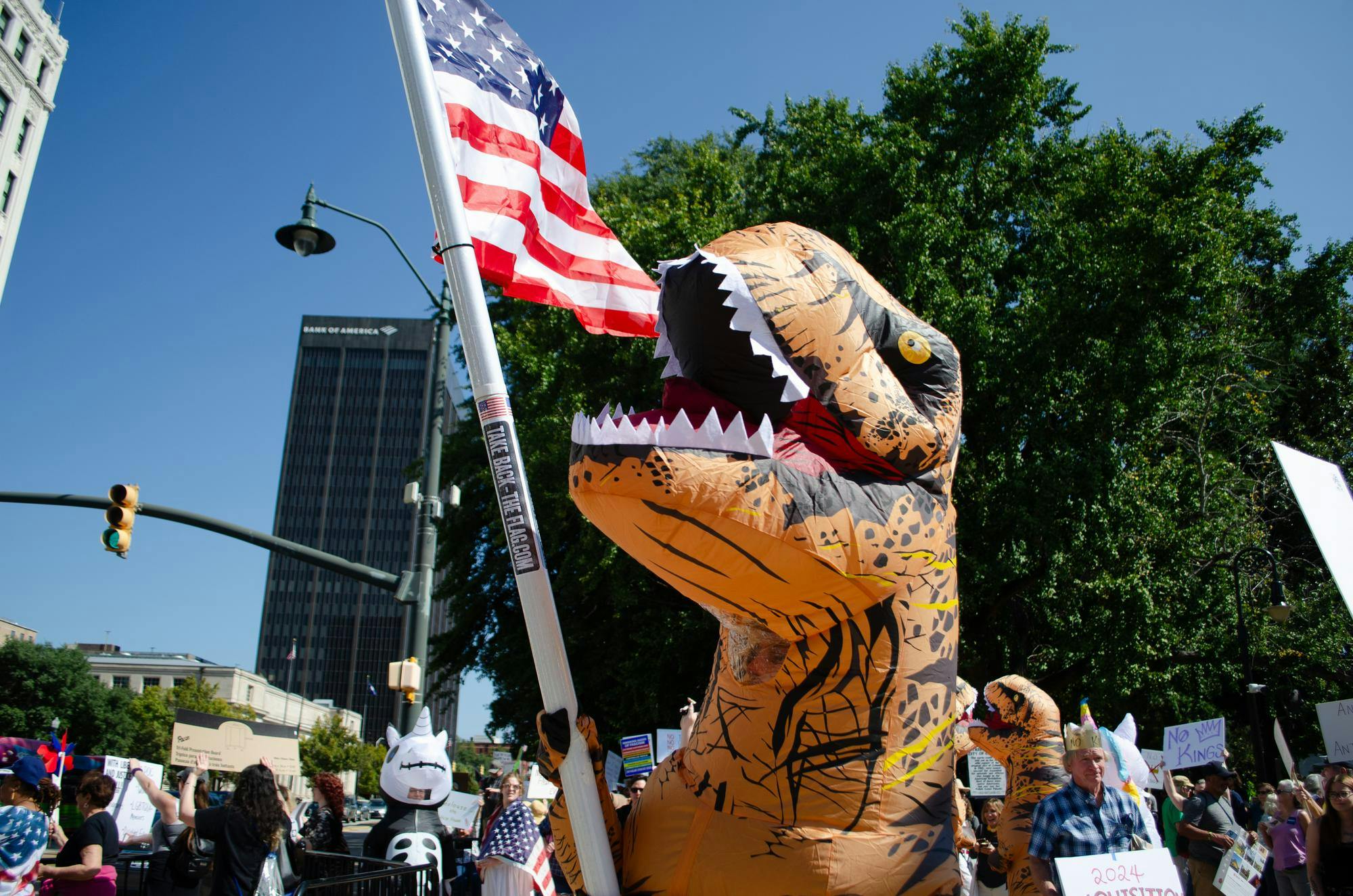 A protester in an inflatable T. Rex costume waves an American flag at a "No Kings" protest in Columbia, South Carolina on Oct. 18, 2025. Other protestors wearing inflatable costumes can be seen in the background.