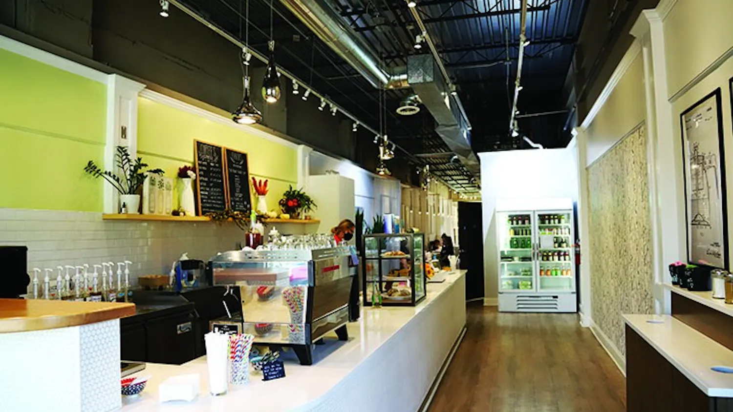 Blum Coffee's bar is brightly lit and located in the front of the store. The bar includes two large chalkboard menus and is lined with coffee machines, syrups and a case of baked goods.