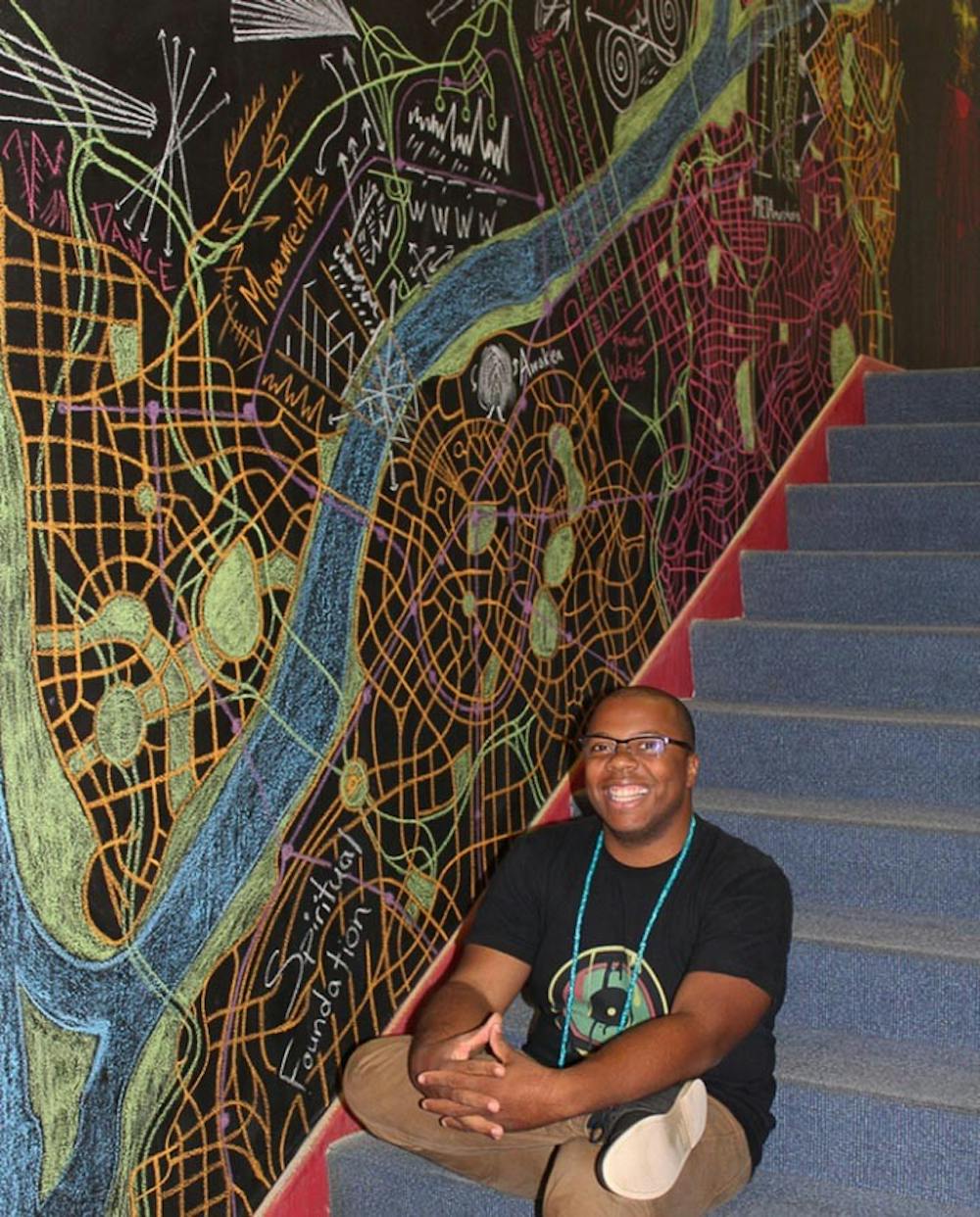 <p>Michael Dantzler sits next to his mural, “Transcend Map,” which is a temporary mural he made in October 2016 for the University of South Carolina School of Music’s library.</p>