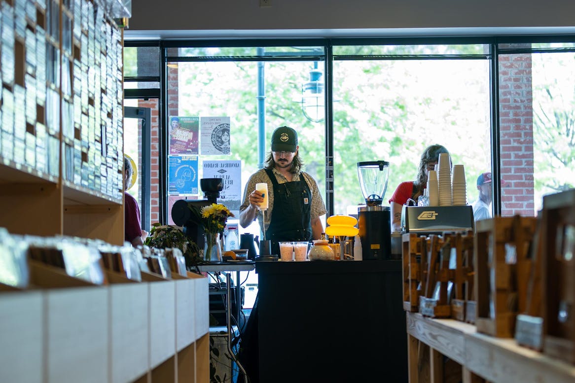 A male barista stands behind the counter for Godspeed Coffee inside the Papa Jazz Record Shoppe preparing a customer's order. He is wearing a green baseball cap, a patterned short sleeve button down shirt, and and apron. Shelves of vinyl records line the aisle leading to the counter.