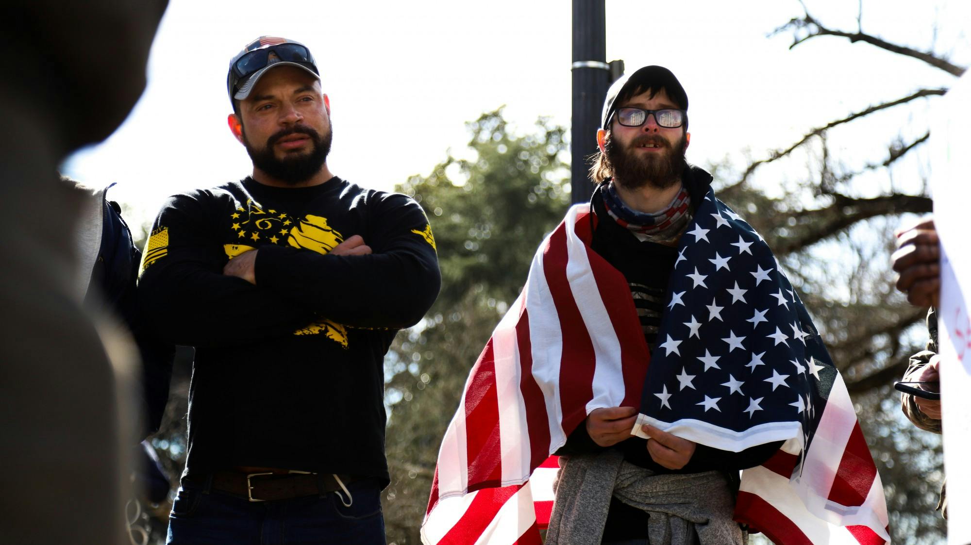 Two “Drive4America” caravan attendees stand side by side watching conversation between other protesters. One of the attendees wore an American flag around his shoulders.&nbsp;