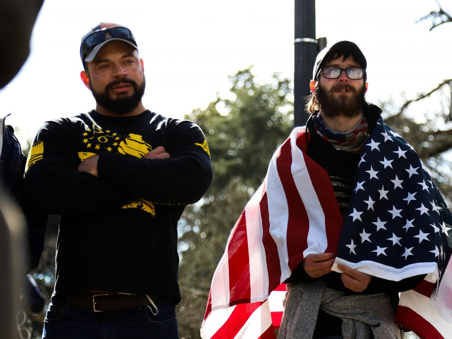 Two “Drive4America” caravan attendees stand side by side watching conversation between other protesters. One of the attendees wore an American flag around his shoulders.&nbsp;
