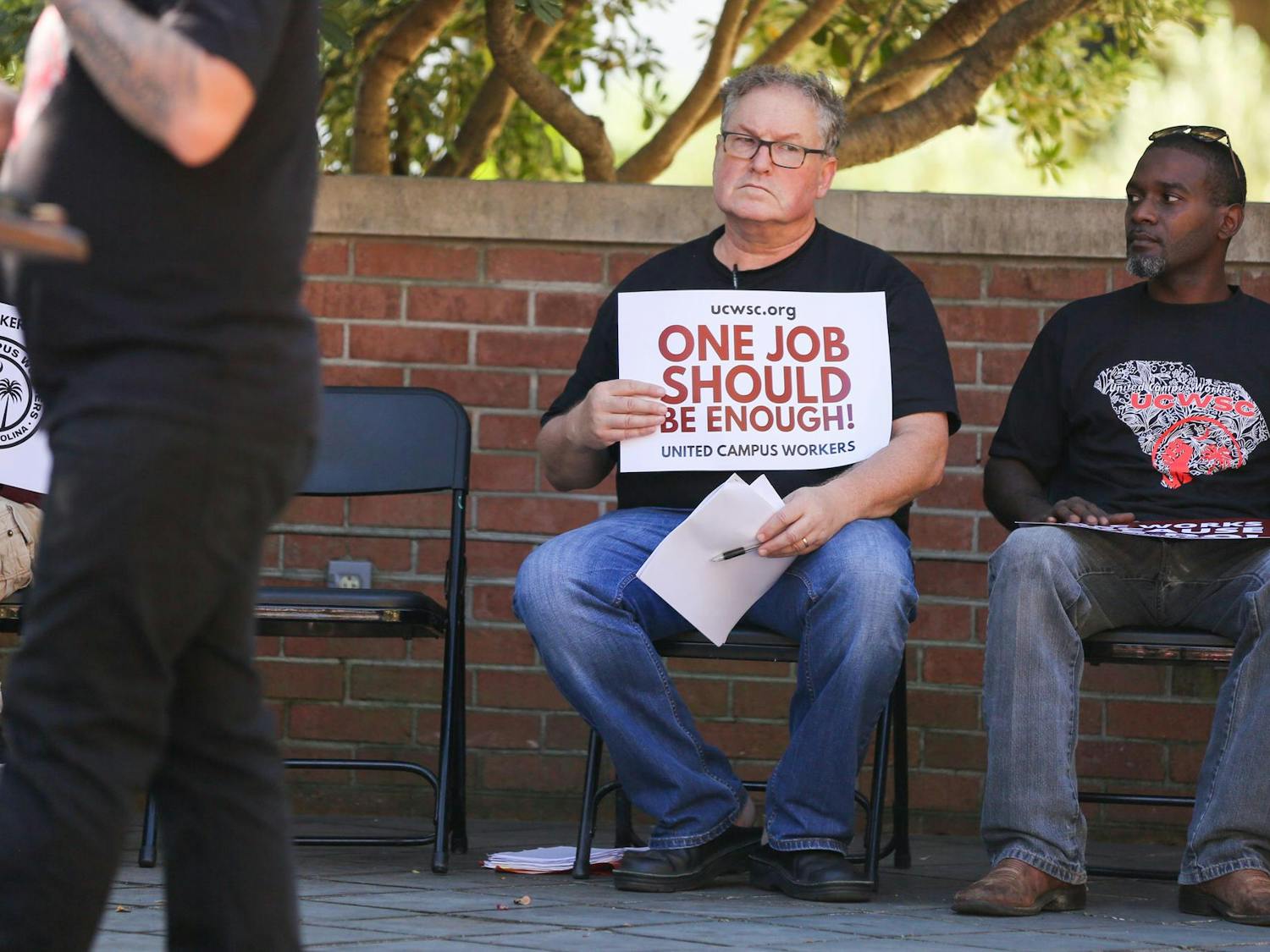 Thomas Alexander, an employee with the USC's department of languages, literatures and cultures, holds a sign stating 'One job should be enough,' while he sits and listens to a colleague speak during a USC Worker Speak-Out event held on the Russell House patio on Oct. 26, 2023. The event was hosted by United Campus Workers at USC.