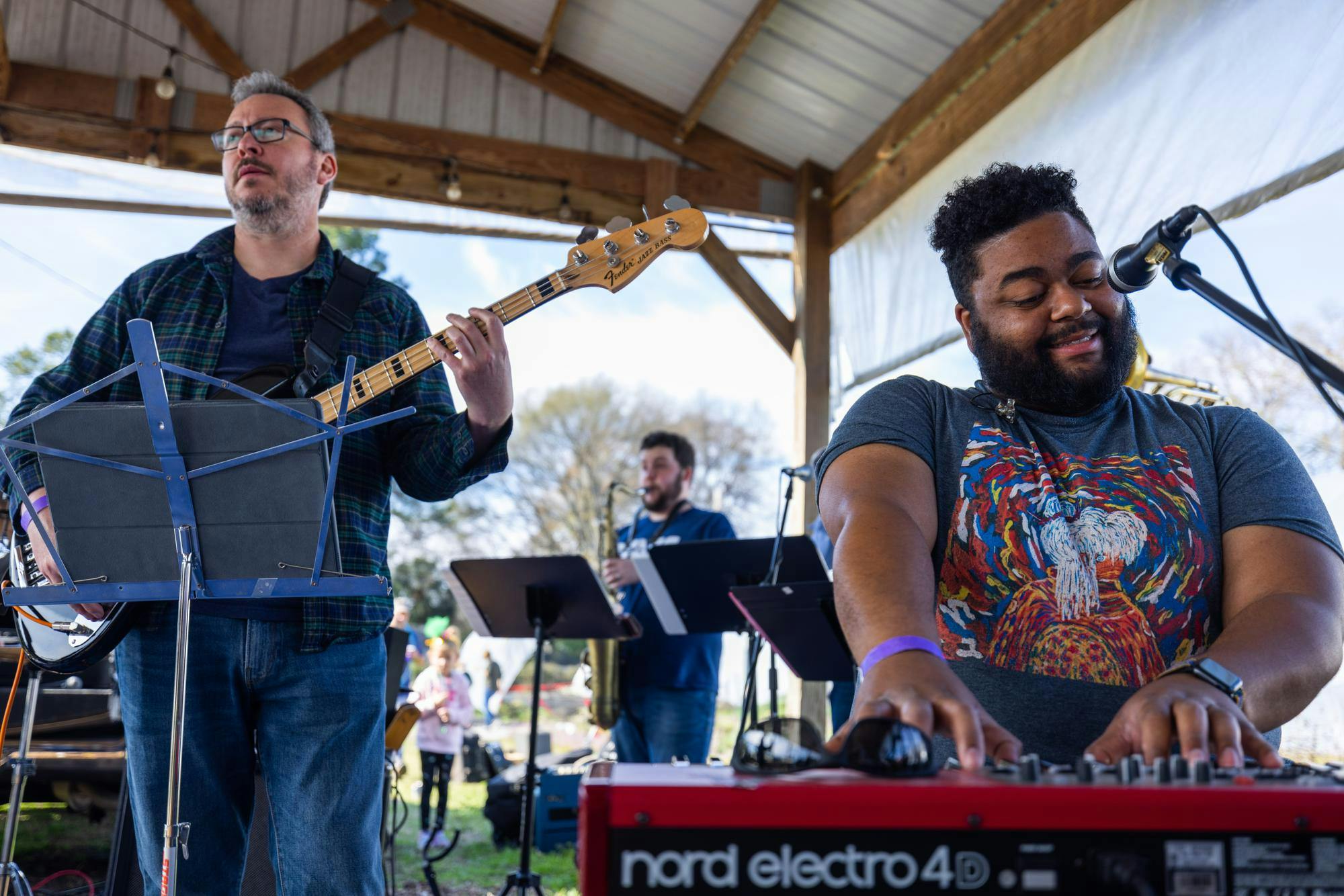 Two musicians perform under an outdoor pavilion, one playing bass guitar and the other smiling while playing a red keyboard, with microphones and music stands set up around them.