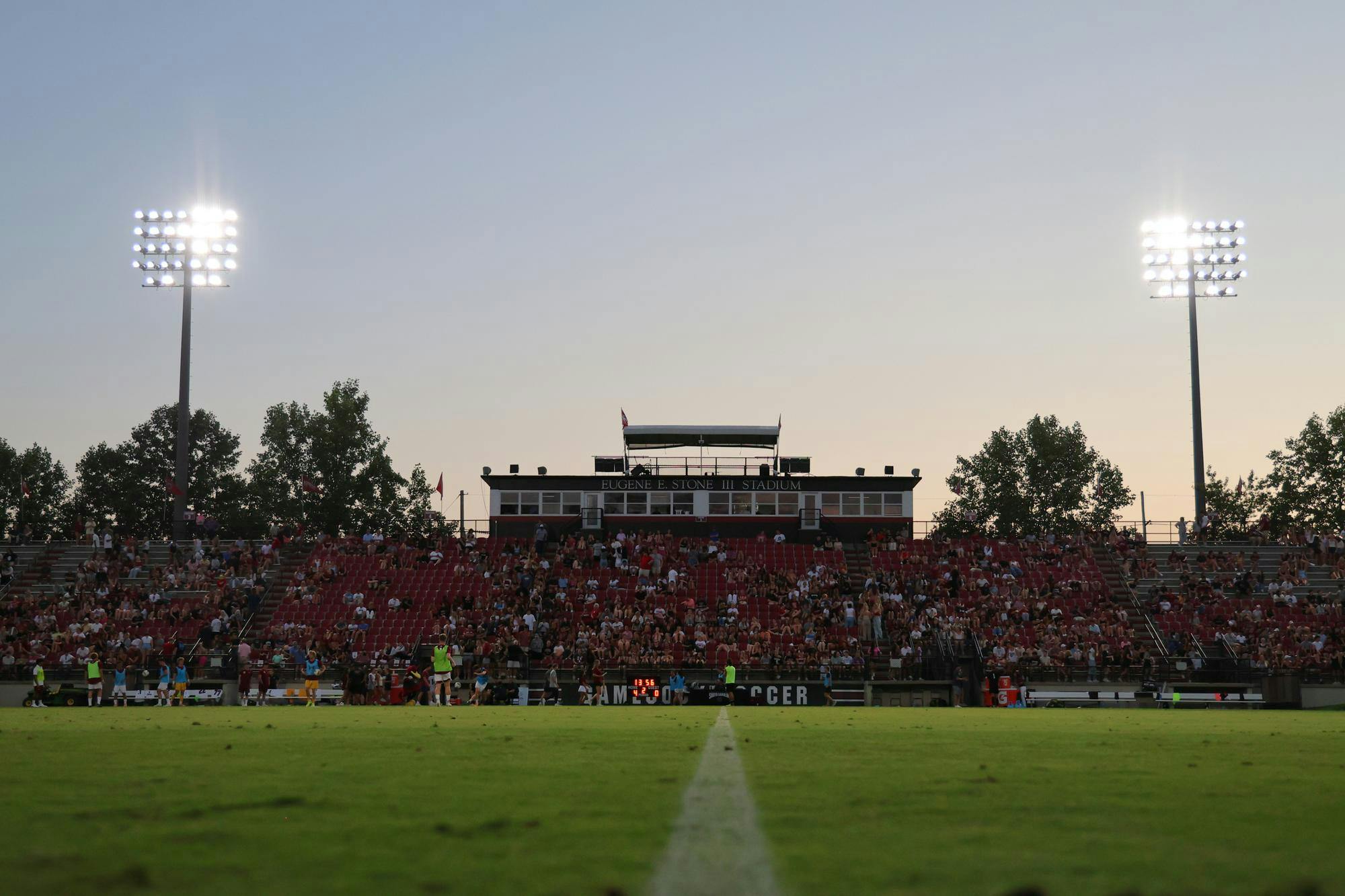 A stadium is pictured with red seats, two light poles on either end of the image and a press box that says "Eugene E. Stone III Stadium" on top. The lights are on and there are fans in the seats.