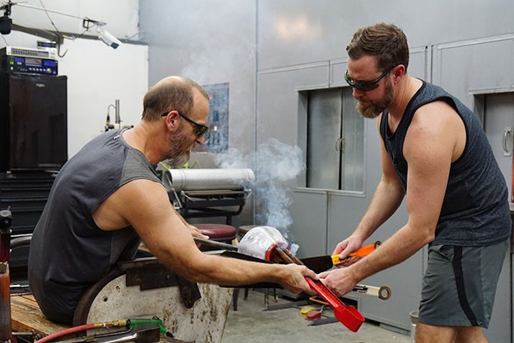 Tom Lockart and Ryan Crabtree open the top of their molded glass to prepare it to be flattened into a plate while smoke rises. The duo could be heard listening to a playlist of '90s rock while they worked, including the Red Hot Chili Peppers' "Higher Ground."