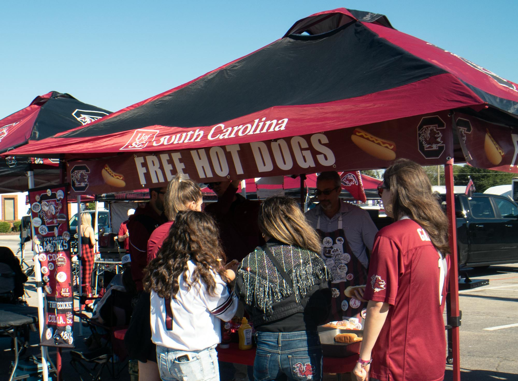 A man wearing a grey polo shirt and black apron serves food to college students wearing red, black and white fan gear prior to a college football game. 