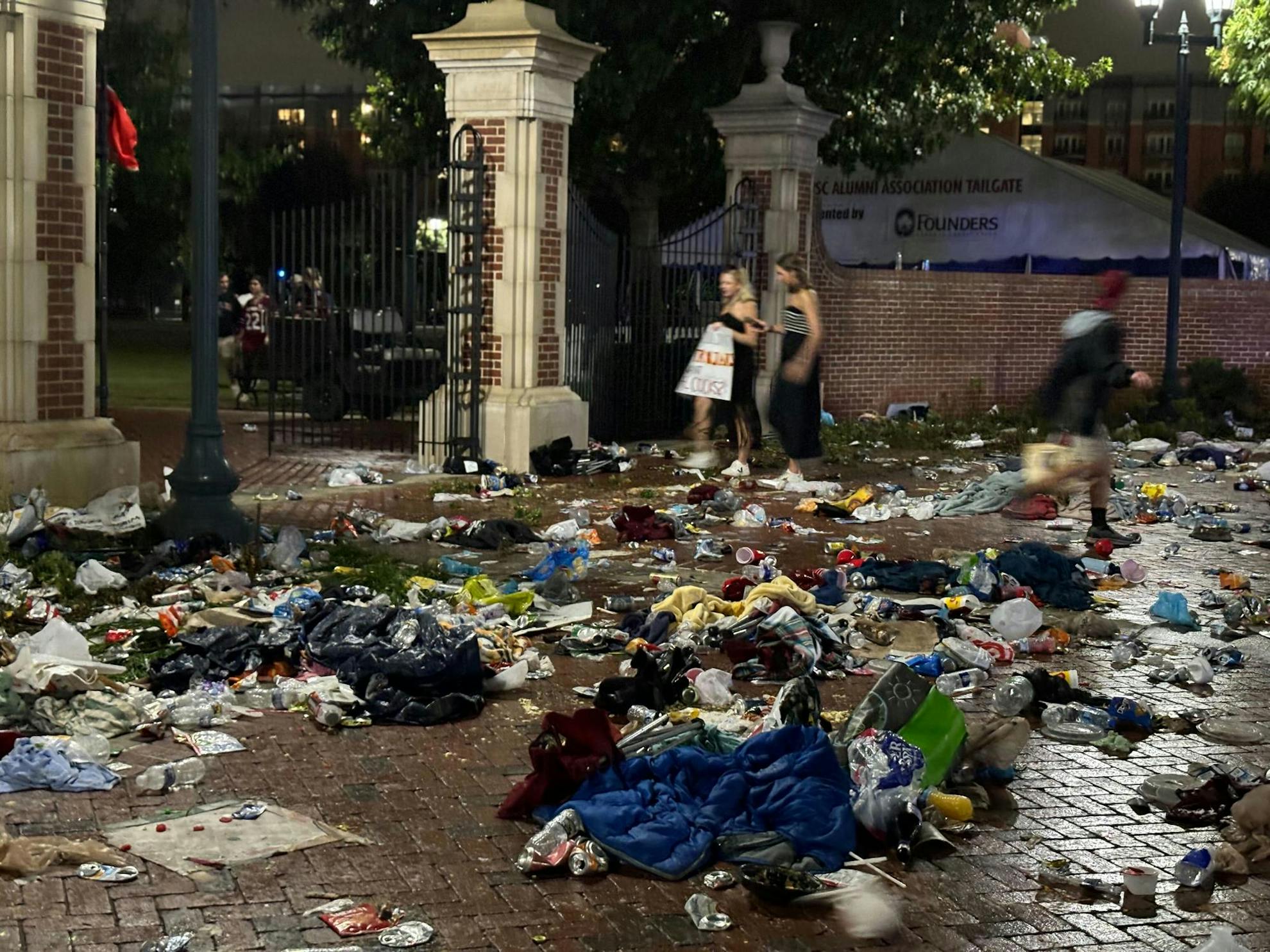 Piles of trash, including bags, blankets and water bottles, are left all over a sidewalk. Three people can be seen walking in the background, two entering the gates to Gamecock Park and one leaving. 