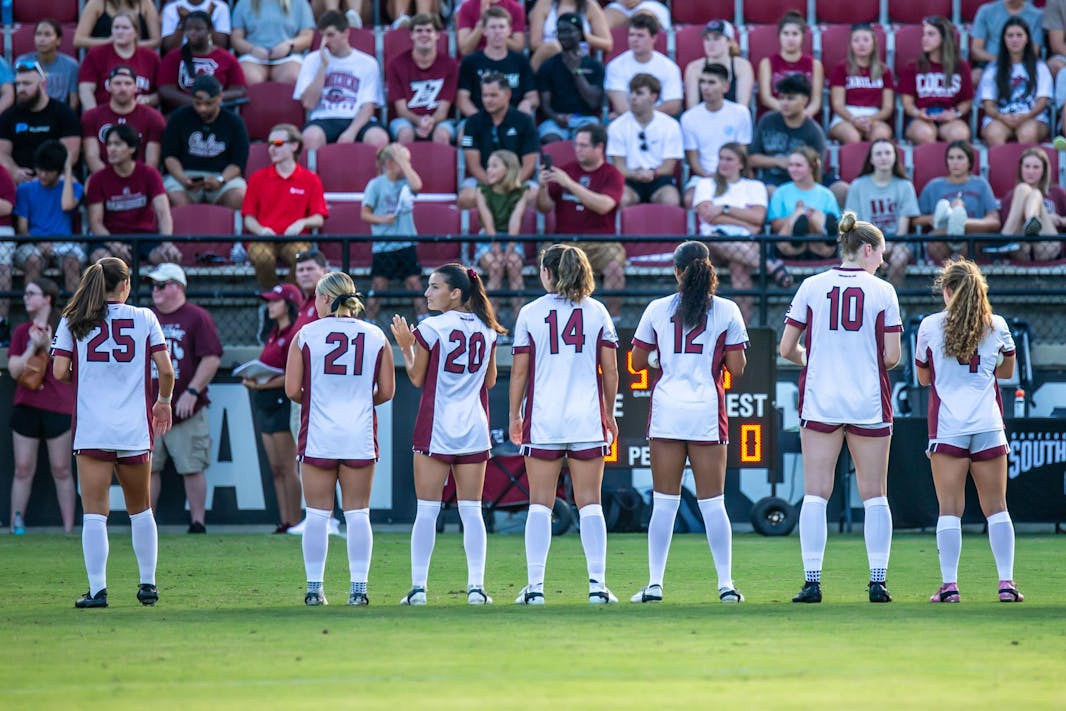 Seven South Carolina women's soccer players, wearing white and red uniforms, stand lined up side-by-side on the side line of a soccer pitch. Their backs are shown as they are facing toward fans in the grandstand. 