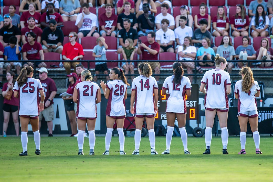 Seven South Carolina women's soccer players, wearing white and red uniforms, stand lined up side-by-side on the side line of a soccer pitch. Their backs are shown as they are facing toward fans in the grandstand.