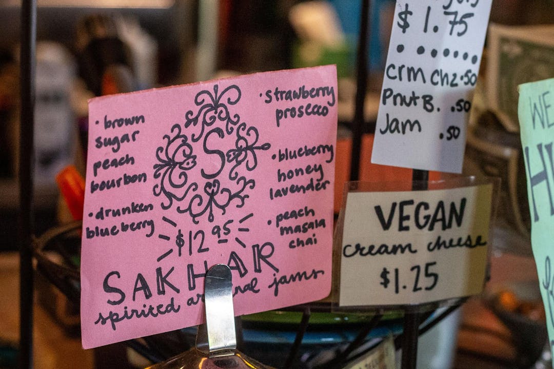 A pink sign sits on the wooden counter of a coffee shop. The sign reads “Sahkar Spirited Artisanal Jam” with the logo hand drawn in the center and flavors, such as brown sugar peach bourbon, drunken blueberry, strawberry prossecco, blueberry honey lavendar and peach masala chai listed around it. 