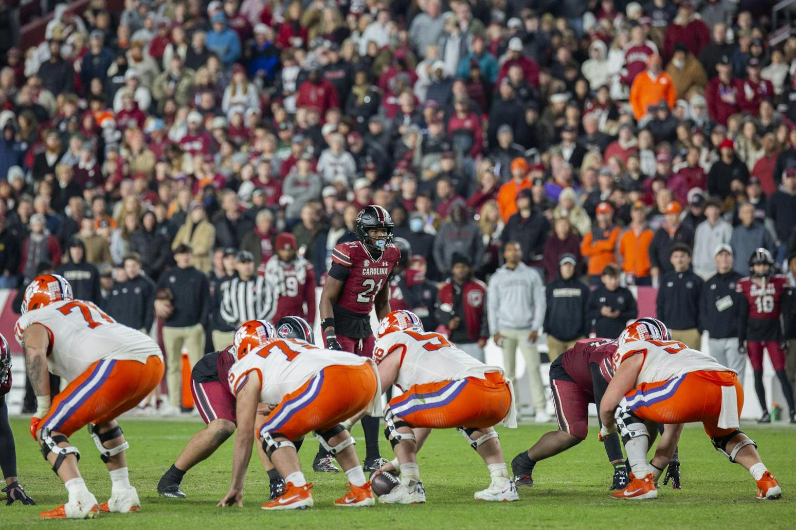 A South Carolina football player stands, looking down on the Clemson offense who is crouched in preparation for the coming play. In the background, spectators stand, watching from the stands. 